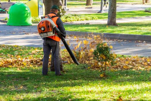 a male worker uses a leaf blower to remove leaves in a park in autumn tidying up and preparing the garden for winter photo